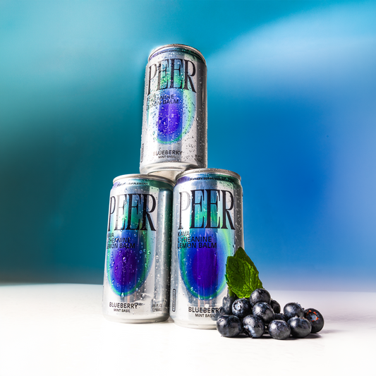 Three silver PEER can drinks with blueberry-infused label graphics are stacked in a pyramid on a white surface, with blueberries and mint leaves placed in front, against a blue gradient background.
