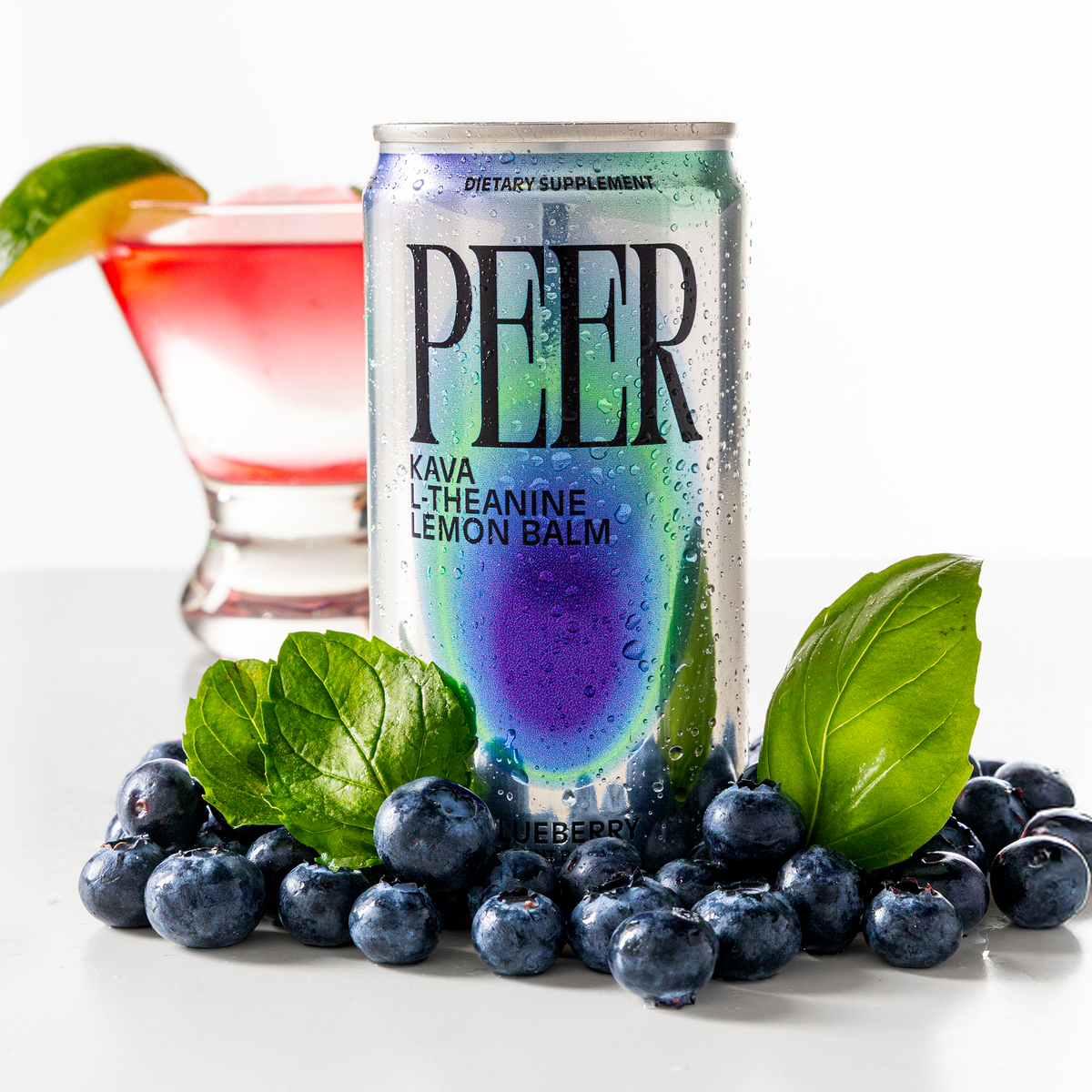 A can of PEER Kava L-Theanine Lemon Balm dietary supplement, styled with fresh blueberries, mint leaves, and a glass of pink drink in the background on a white surface.