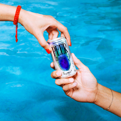 A person's hands holding a can of Peer Blueberry over a bright blue swimming pool background.