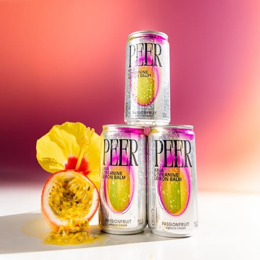 Three silver cans of PEER balm with pink and yellow graphics are arranged in a pyramid on a white surface, with a cut passionfruit and a yellow flower beside them against a pink-to-orange gradient background.