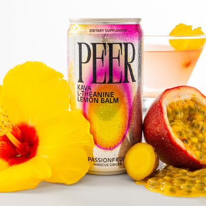 A can of PEER Kava L-Theanine Lemon Balm dietary supplement stands on a white surface, surrounded by tropical ingredients including a yellow hibiscus flower, a passionfruit half, fresh ginger, and a pink drink in a glass with a passionfruit garnish.