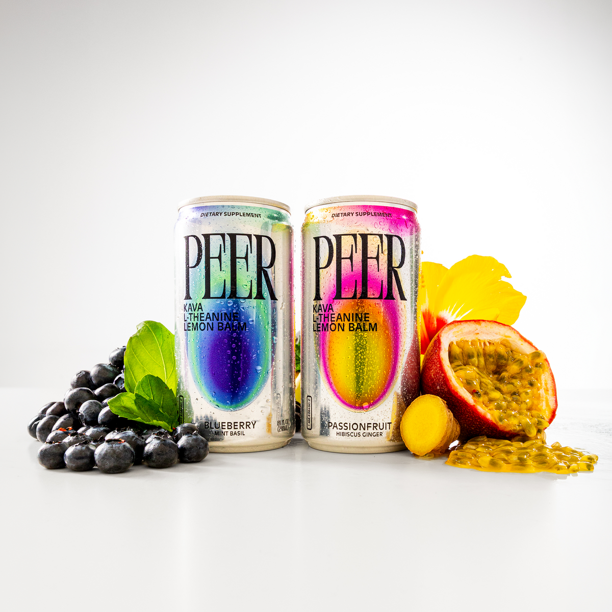 Two tall cans of PEER Kava L-theanine lemon balm drink are shown on a clean white studio background, styled with blueberries, mint leaves, a passionfruit half, and yellow citrus pieces. The left can is blueberry flavor and the right can is passionfruit flavor.